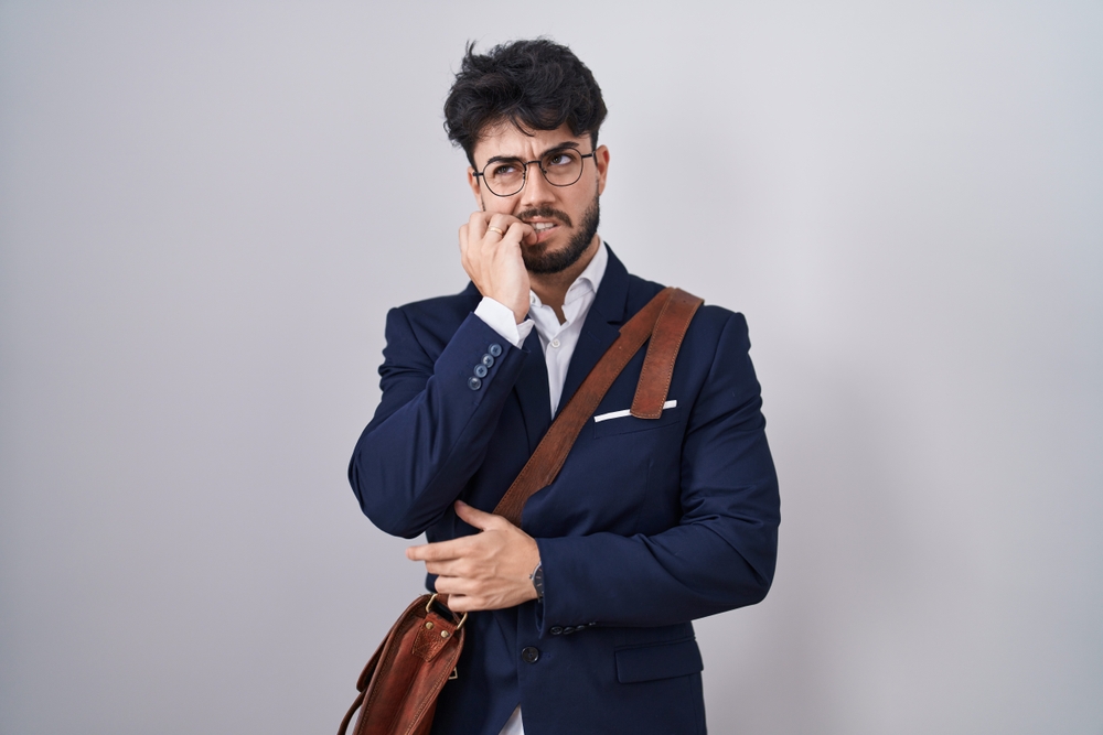 Hispanic man with beard wearing business clothes looking stressed and nervous with hands on mouth biting nails. anxiety problem.