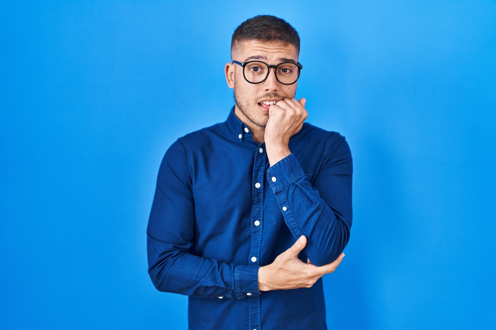 Young hispanic man wearing glasses over blue background looking stressed and nervous with hands on mouth biting nails. anxiety problem.